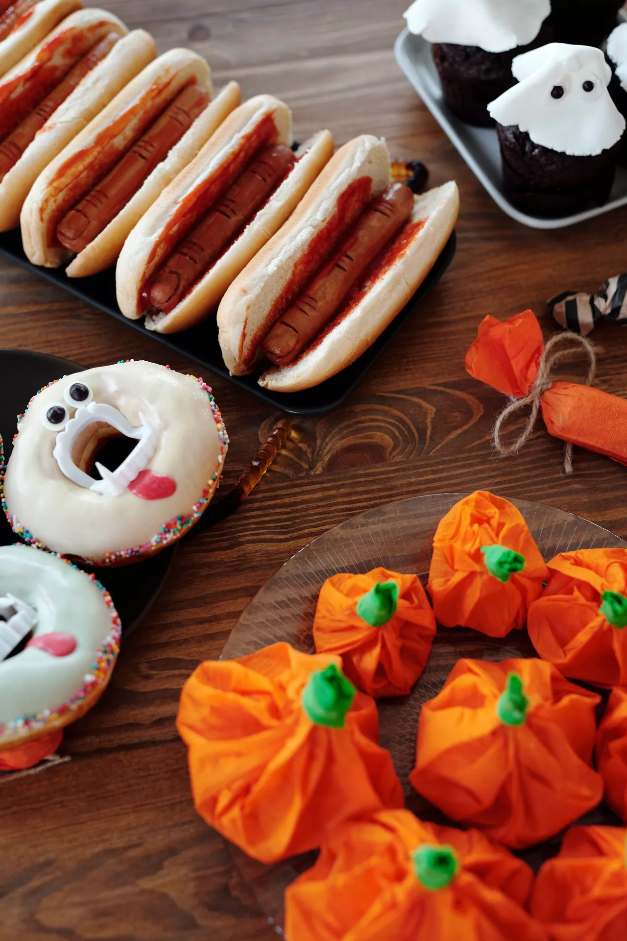 Halloween Party Food Ideas featuring Hot Dogs, Monster Donuts & Pumpkin Goodie Bags, with spooky finger-style hot dogs, ghost cupcakes, monster donuts, and bright orange pumpkin-shaped treat bags arranged on a festive Halloween party table.