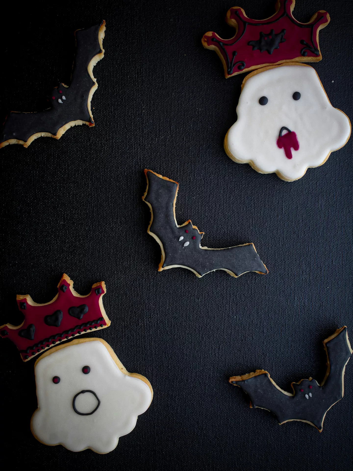 Halloween Party Food Ideas featuring ghost and bat cookies decorated with black, white, and burgundy icing, including playful ghost cookies wearing crowns and spooky bat-shaped cookies arranged on a dark background.