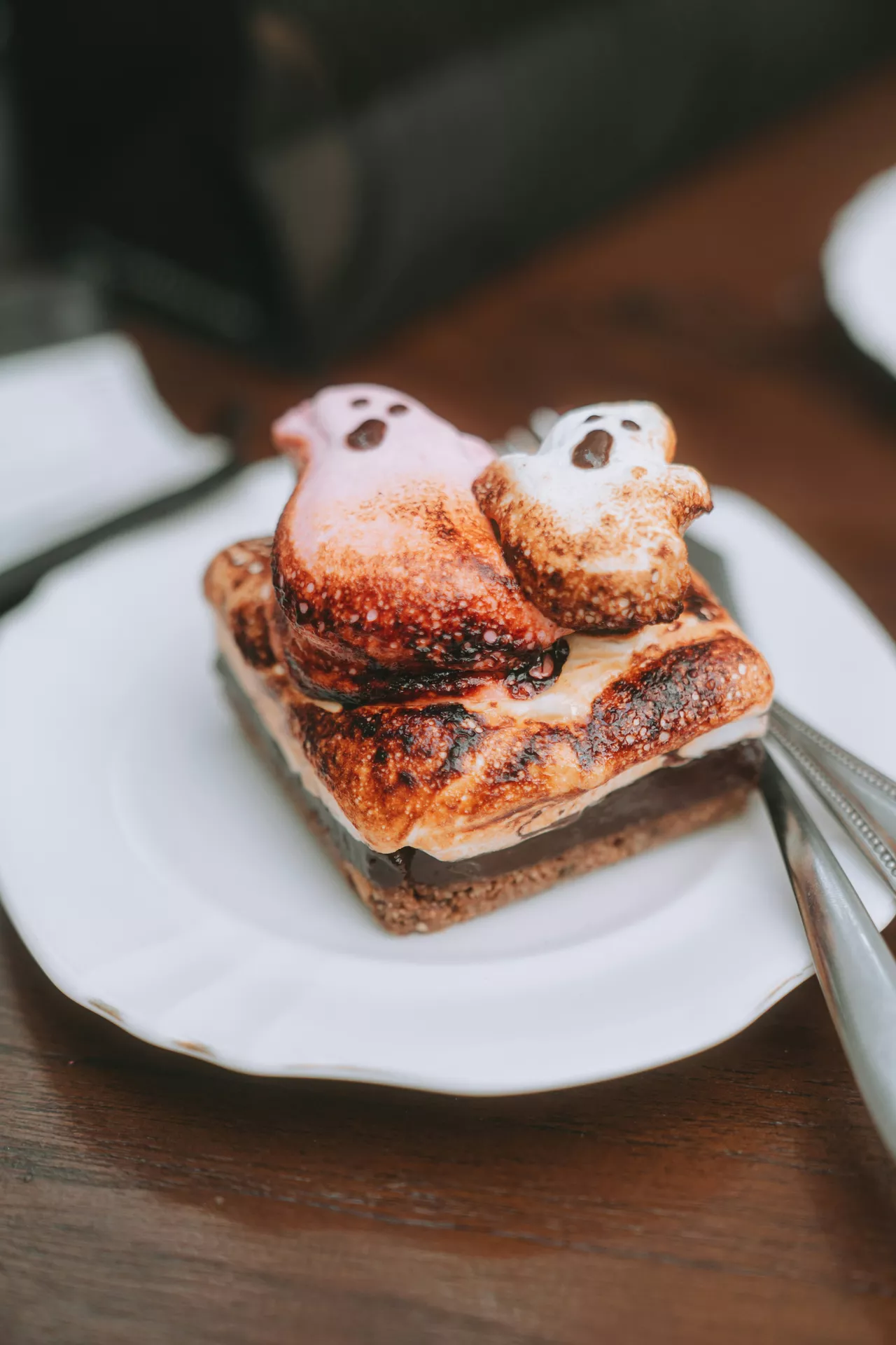 Halloween Party Food Ideas featuring ghostly s'mores made with toasted marshmallows, melted chocolate, graham crackers, and adorable ghost-shaped marshmallow toppers served on a white plate for a spooky Halloween dessert.