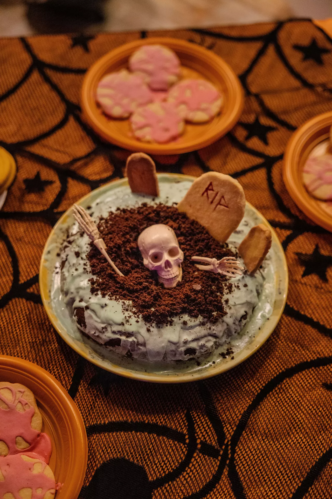 Halloween Party Food Ideas featuring a spooky graveyard cake decorated with chocolate cookie crumbs, edible tombstones, a skeleton skull and hands, surrounded by Halloween cookies on an orange spiderweb tablecloth for a fun party dessert display.
