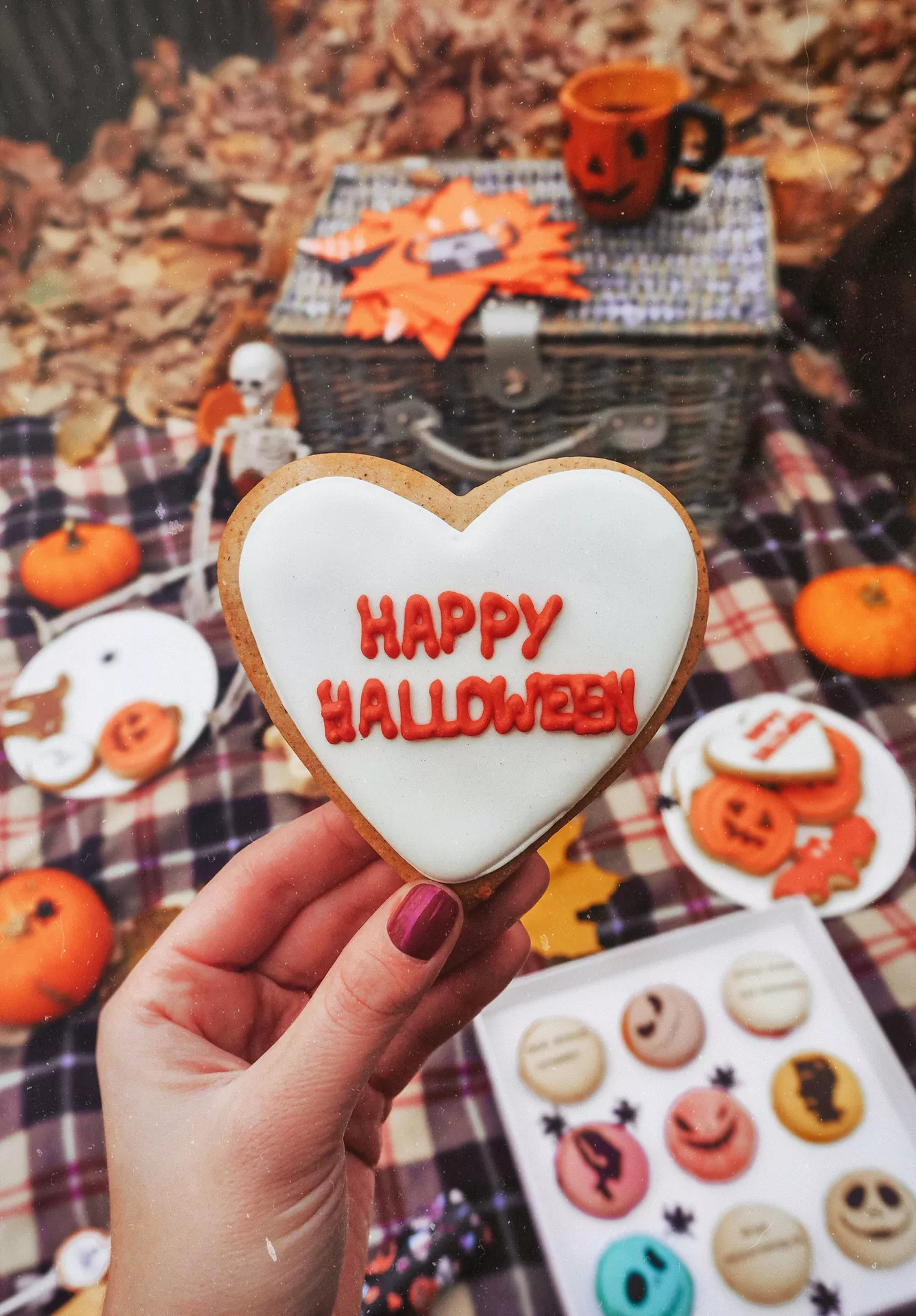 Halloween Party Food Ideas featuring happy halloween cookies, with a heart-shaped iced cookie reading Happy Halloween held in the foreground, surrounded by spooky macarons, pumpkin cookies, mini pumpkins, and a festive Halloween picnic setup.