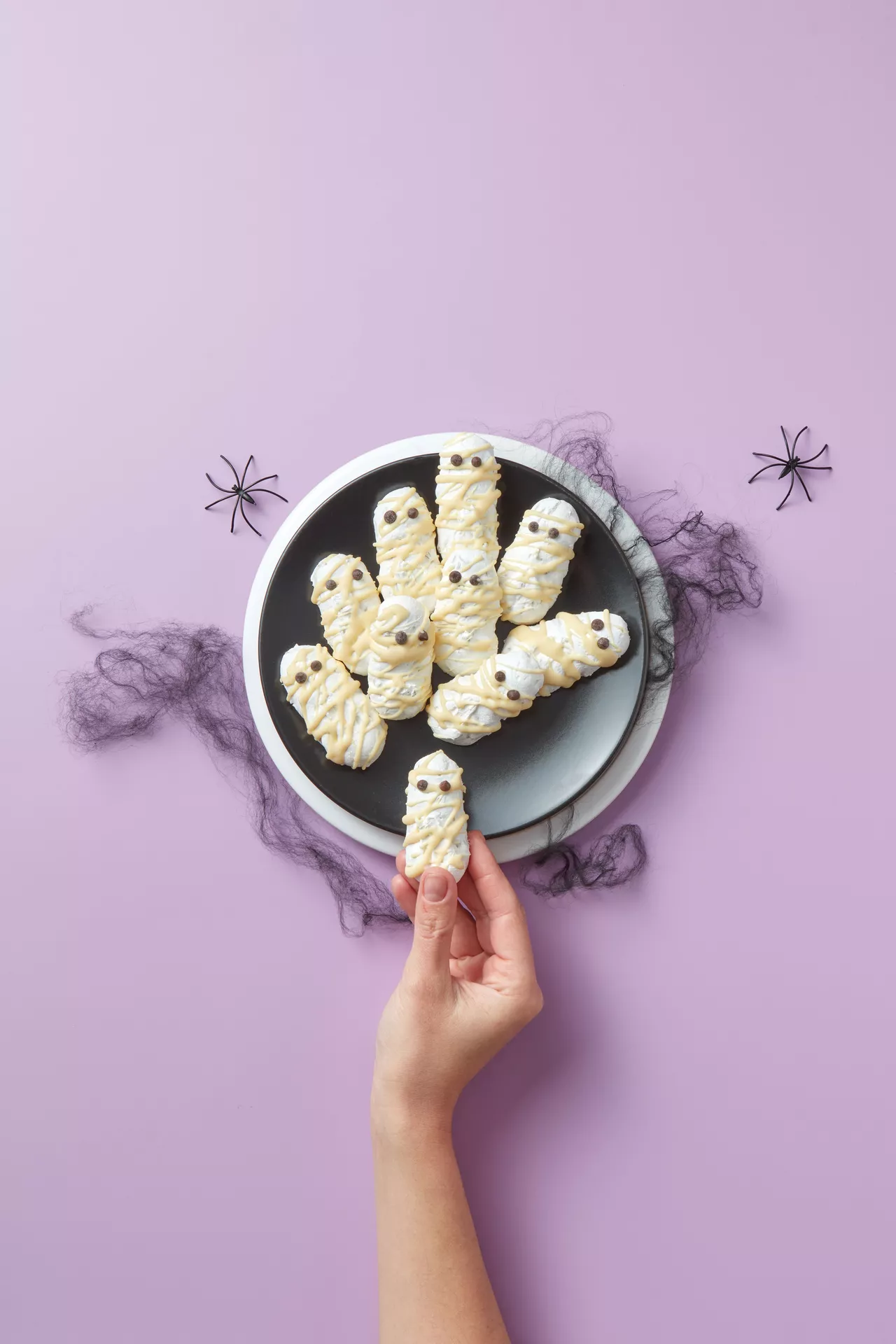 Halloween Party Food Ideas featuring adorable mummy cookies decorated with white chocolate drizzle and candy eyes, arranged on a black plate with spooky spider decorations while a hand holds one cookie against a pastel purple background.