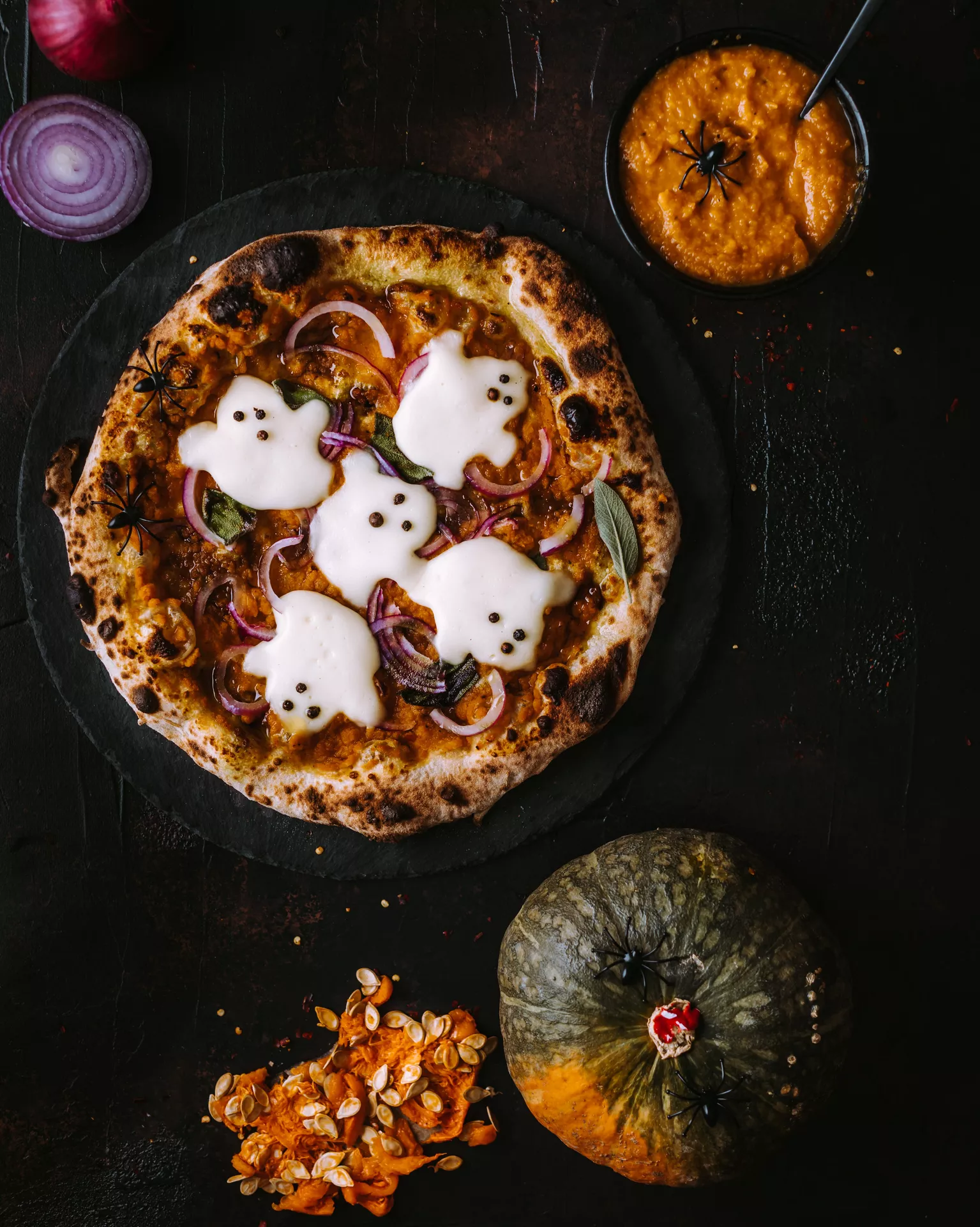 Halloween Party Food Ideas featuring a spooky pizza with mozzarella ghosts topped with red onion and herbs, served alongside pumpkin dip and seasonal Halloween decorations for a festive dinner spread.