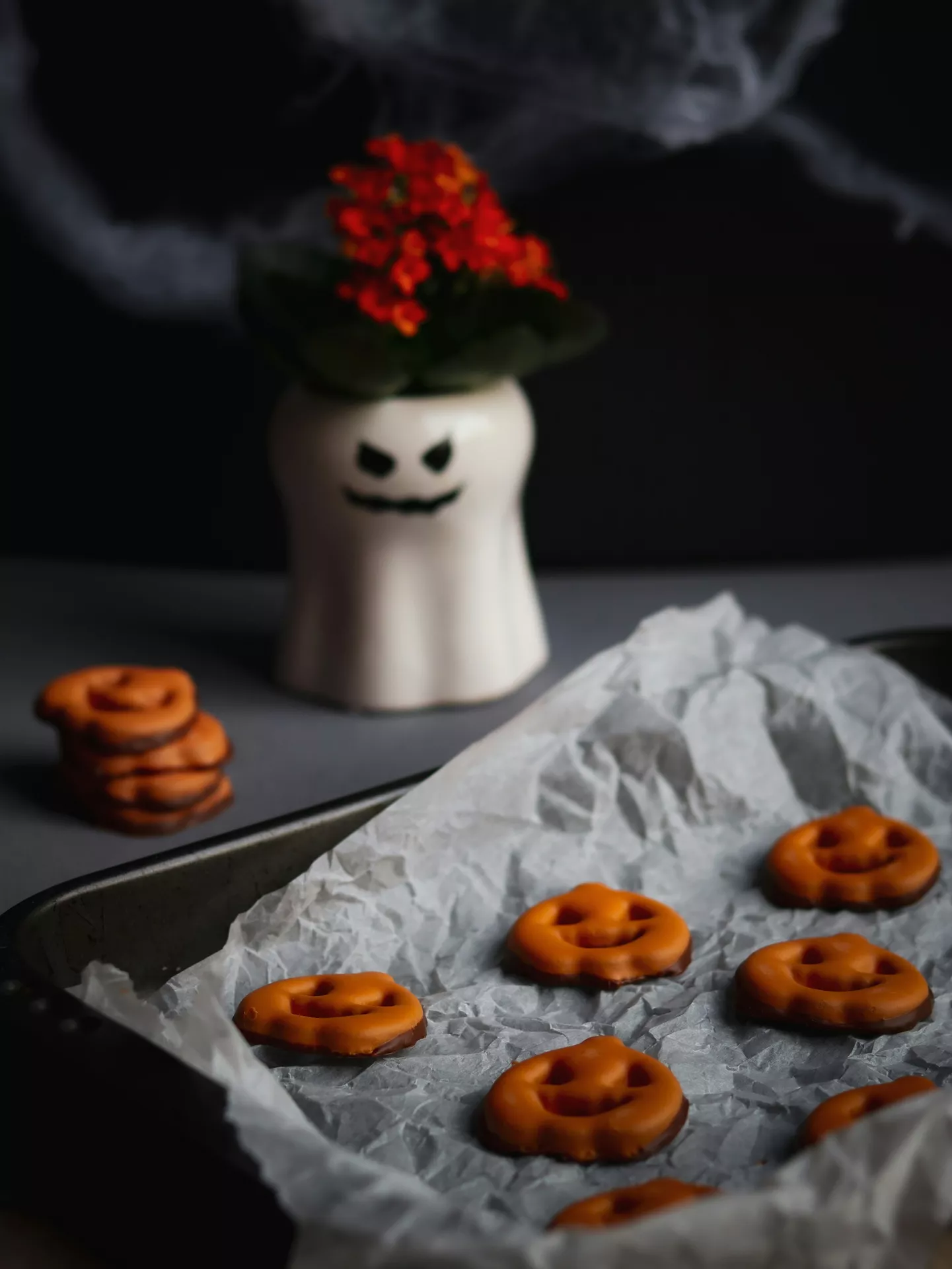 Halloween Party Food Ideas featuring pumpkin cookies made from chocolate-covered pretzels coated in orange candy, arranged on a parchment-lined tray with a playful ghost planter in the background for a festive Halloween treat.