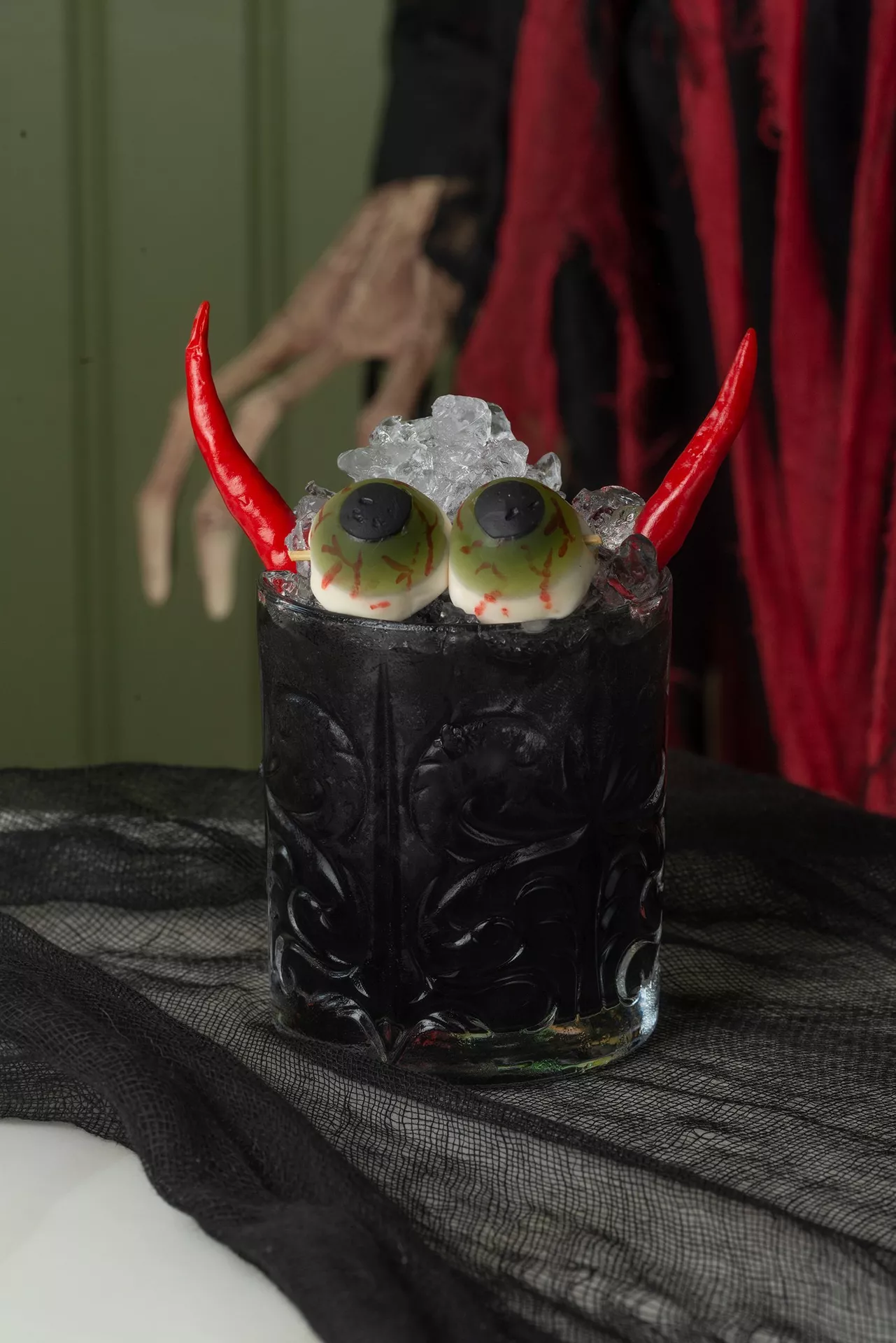 Halloween Party Food Ideas featuring the devil's eyeball cocktail served in a black glass with candy eyeballs, red chili pepper horns, crushed ice, and spooky Halloween decorations in the background.