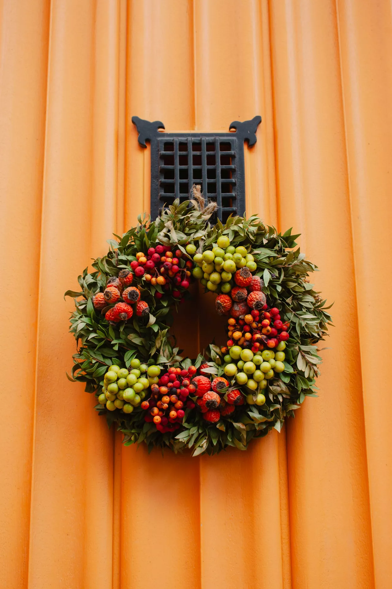 Fall decor wreaths featuring a Colorful Berry Wreath With Rosehips & Green Berries displayed on a warm orange front door, combining lush green foliage, vibrant red rosehips and fresh green berries for a cheerful autumn entrance.