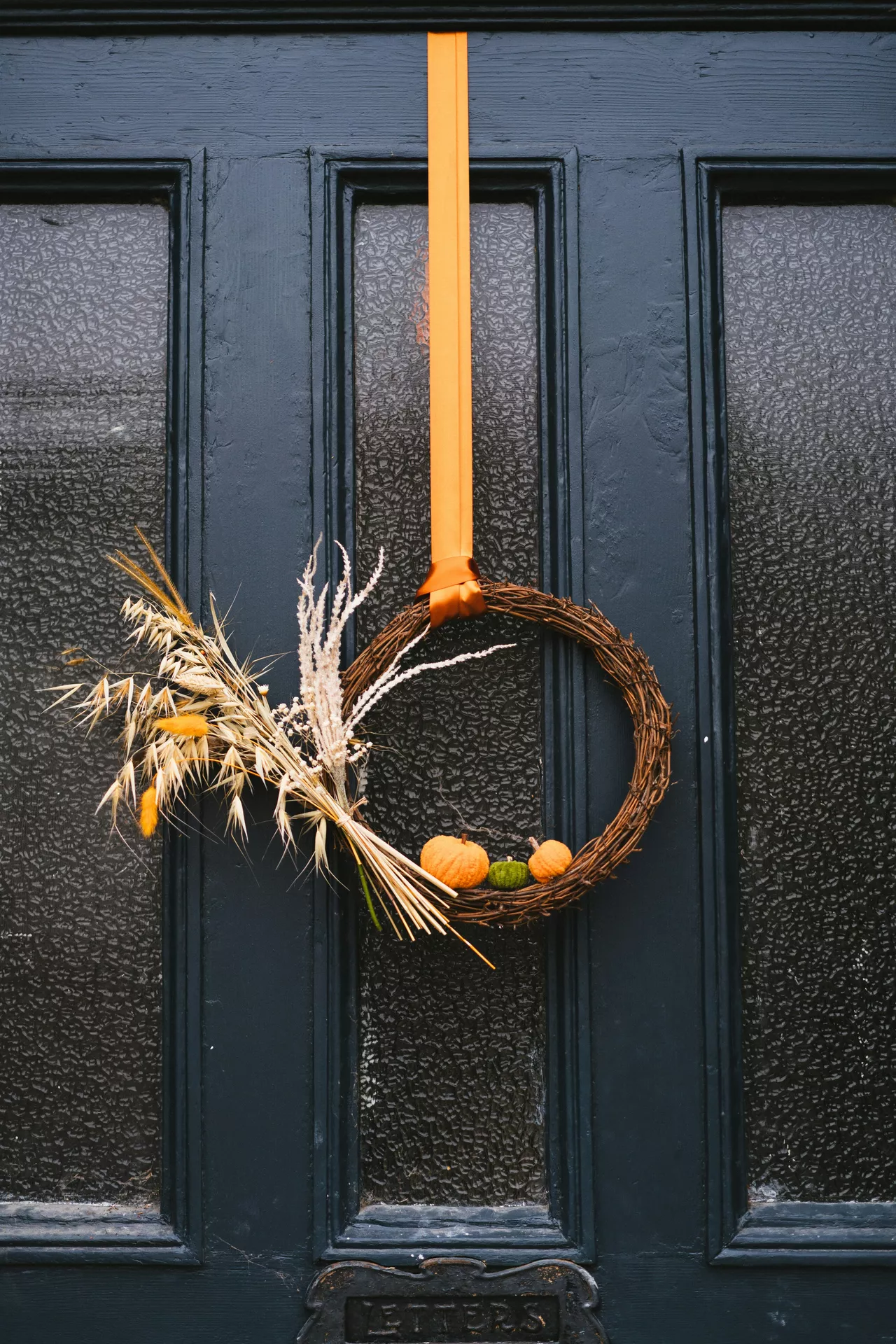 Fall decor wreaths featuring a Grapevine Wreath With Dried Grass Bouquet decorated with natural grapevine, dried grasses, pampas stems and mini pumpkins, hanging from a warm amber ribbon on a black front door for a modern autumn look.