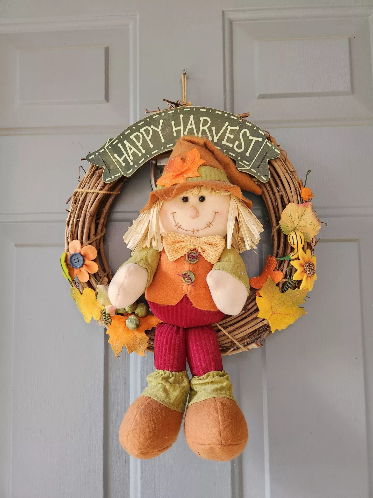 Fall decor wreaths featuring a grapevine wreath with plush scarecrow, decorated with colorful autumn leaves, flowers, and harvest accents, hanging on a gray front door for a cheerful and welcoming seasonal entrance.