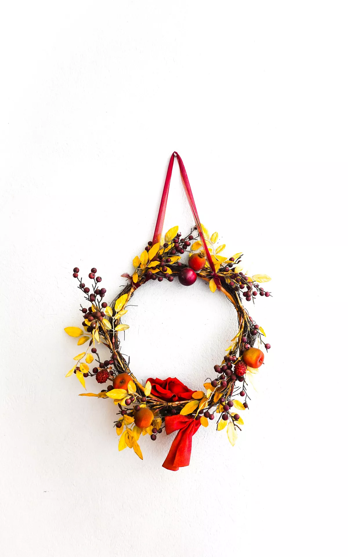 Fall decor wreaths featuring a Minimalist Wreath With Yellow Leaves, Mini Apples & Red Ribbon displayed on a white wall, combining a simple grapevine base with golden leaves, berries, tiny apples and a bright red bow for elegant autumn decor.