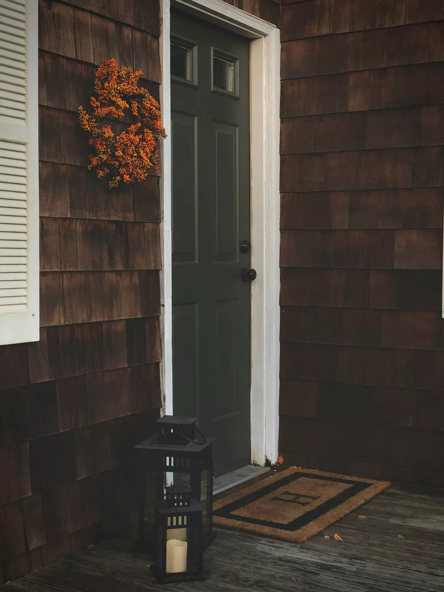 Fall decor wreaths featuring an orange berry wreath displayed beside a dark front door, creating a cozy autumn entryway with warm seasonal colors, a black lantern and a simple welcoming front porch.