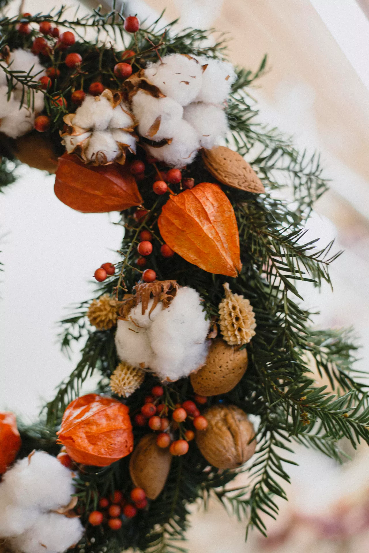 Fall decor wreaths featuring raw cotton balls, everygreens and chinese lanterns arranged with fresh evergreen branches, red berries, walnuts and natural botanicals, creating a cozy rustic wreath full of warm autumn texture and seasonal charm.