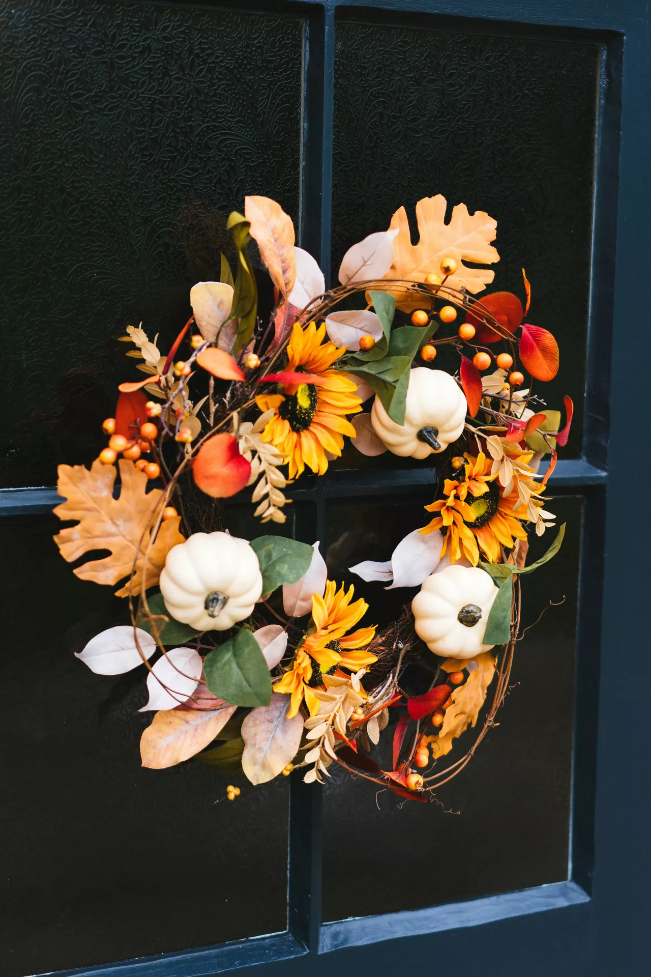 Fall decor wreaths featuring white pumpkins, leaves, sunflowers and berries, arranged on a grapevine wreath with warm autumn foliage and seasonal accents, hanging on a dark front door for a cozy fall entrance.
