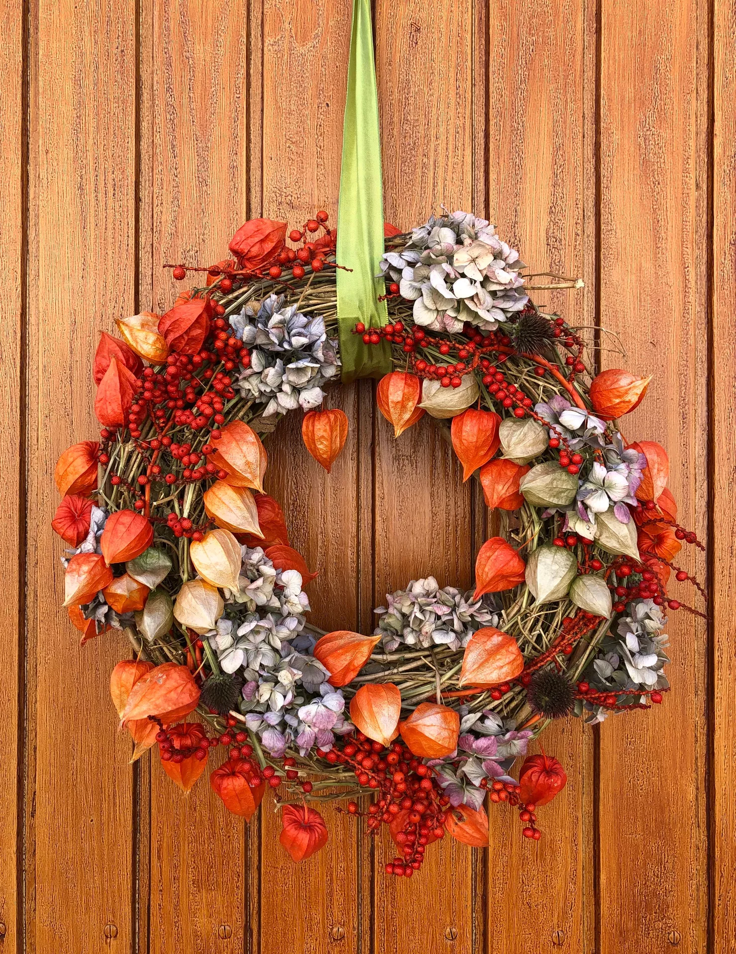 Fall decor wreaths featuring a Wreath With Chinese Lanterns, Physalis & Red Berries, beautifully arranged with dried hydrangeas, vibrant orange lantern pods, and rich red berries, hanging on a wooden door for an elegant autumn entrance.
