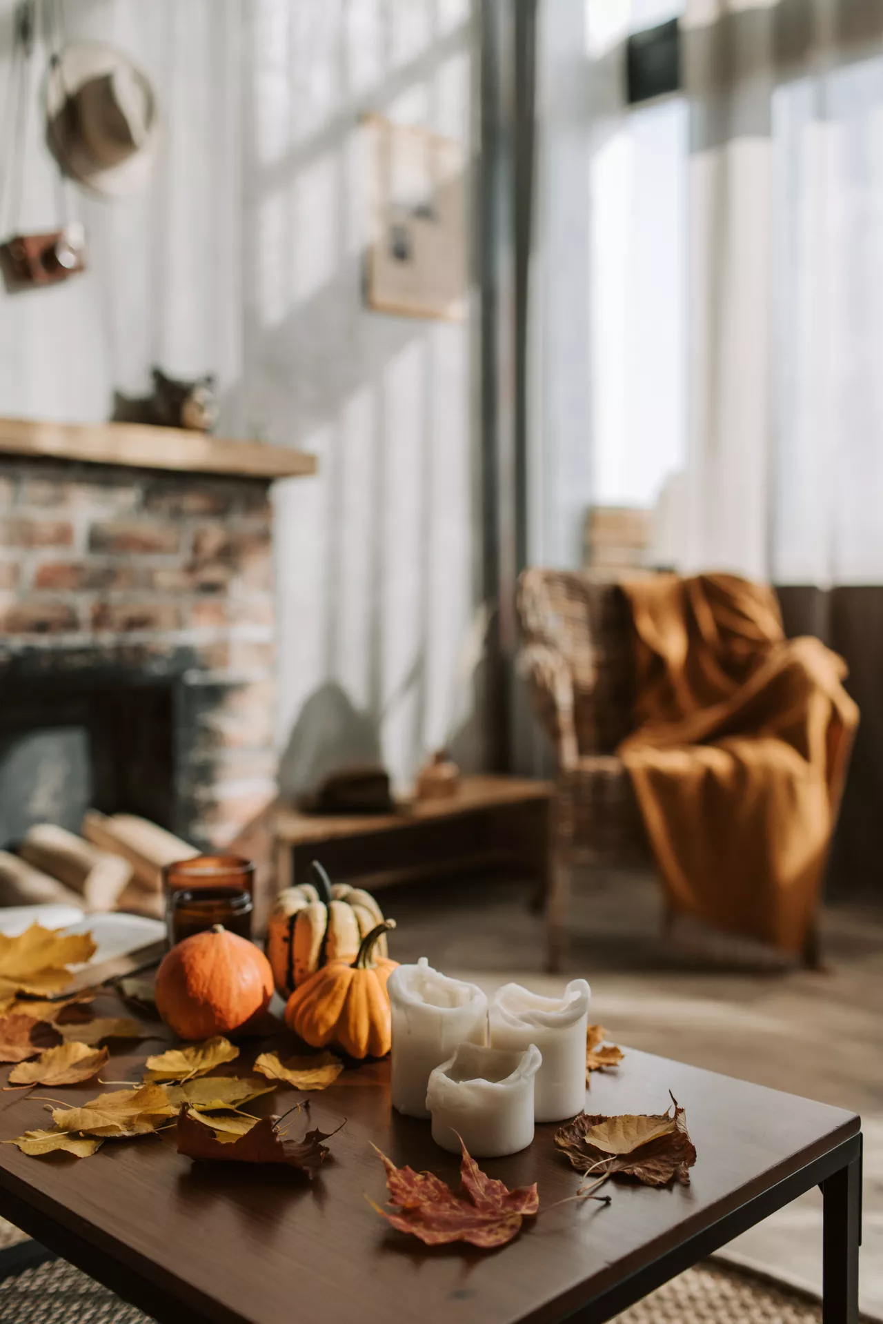Fall living room decor ideas featuring candles, fall leaves and candles on coffee table alongside mini pumpkins, with a cozy fireplace and armchair draped in a warm orange blanket in the background.