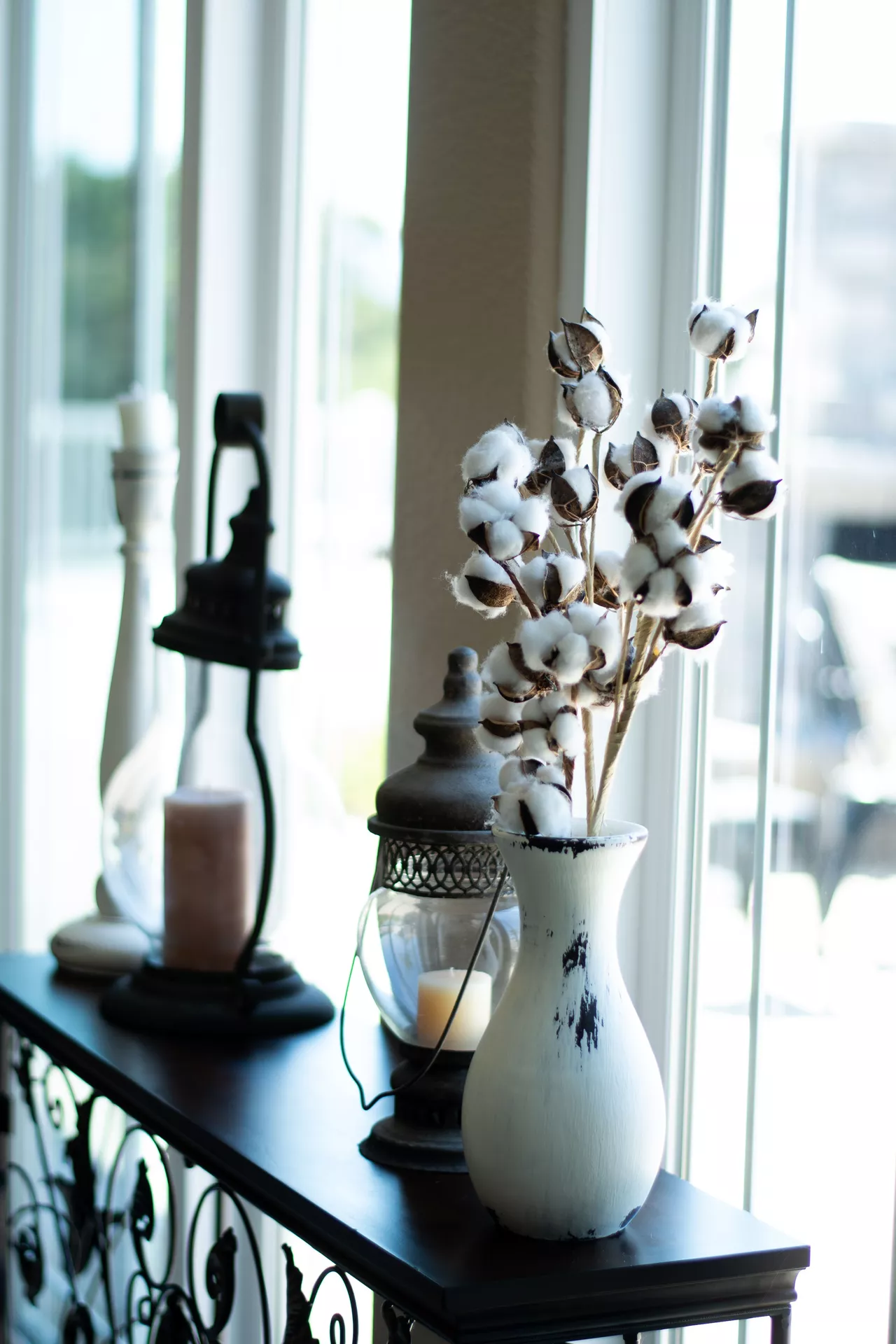 Fall living room decor ideas featuring a cotton plant displayed in a rustic white vase beside vintage-style lanterns and candles on a dark console table by a bright window.