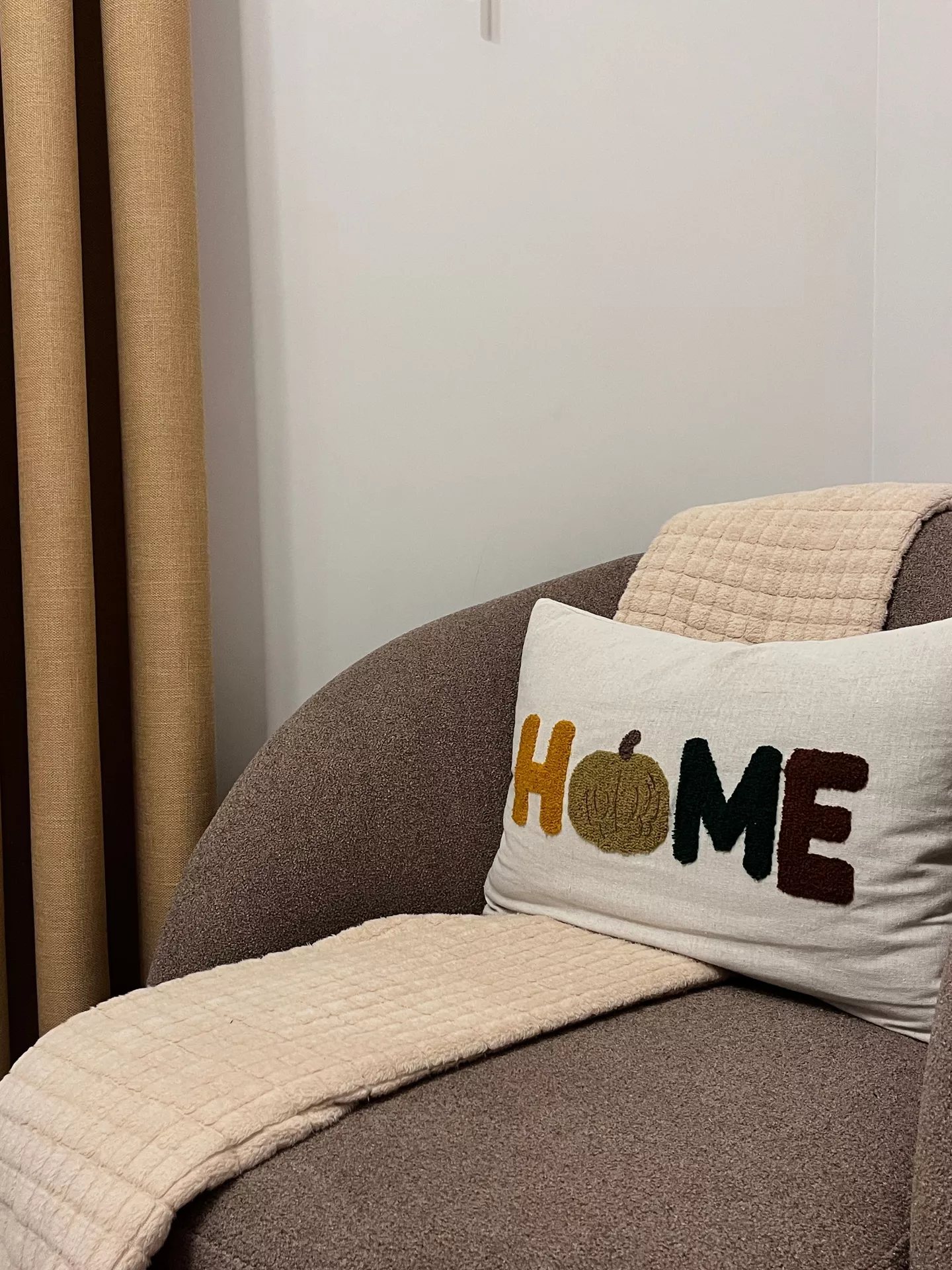 Fall living room decor ideas featuring a cute fall themed cushion with colorful HOME lettering and a pumpkin detail, styled on a cozy brown armchair with soft cream blankets.