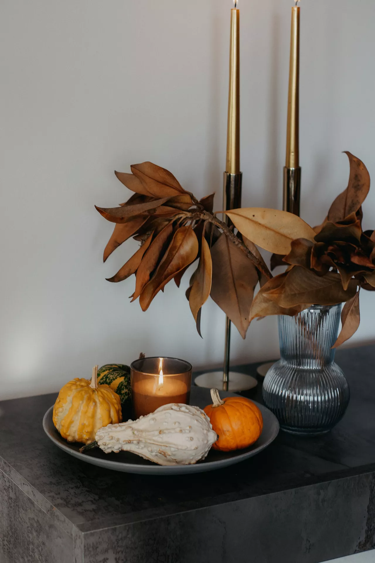 Fall living room decor ideas featuring a mix of pumpkins and candle on a tray, paired with dried autumn leaves in a glass vase and tall gold taper candles for a warm seasonal display.