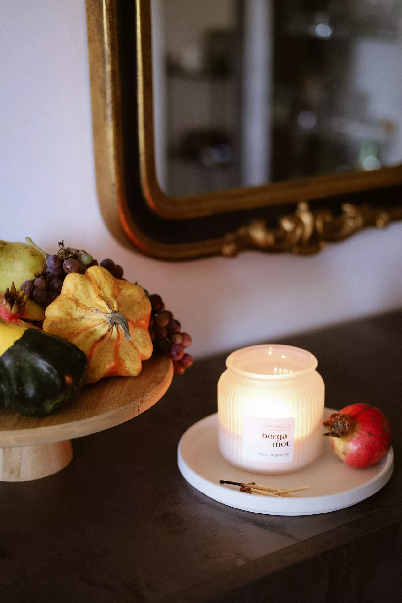 Fall living room decor ideas featuring a glowing scented candle on a white tray beside a pomegranate, with pumpkins, pears, and grapes arranged on a wooden pedestal for a cozy autumn atmosphere.