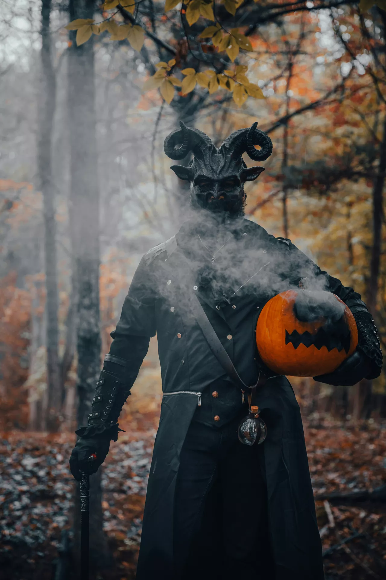 Halloween costume ideas featuring a mysterious black devil dressed in a dark gothic outfit with a horned demon mask, black gloves, and a smoking jack-o-lantern in a misty autumn forest.