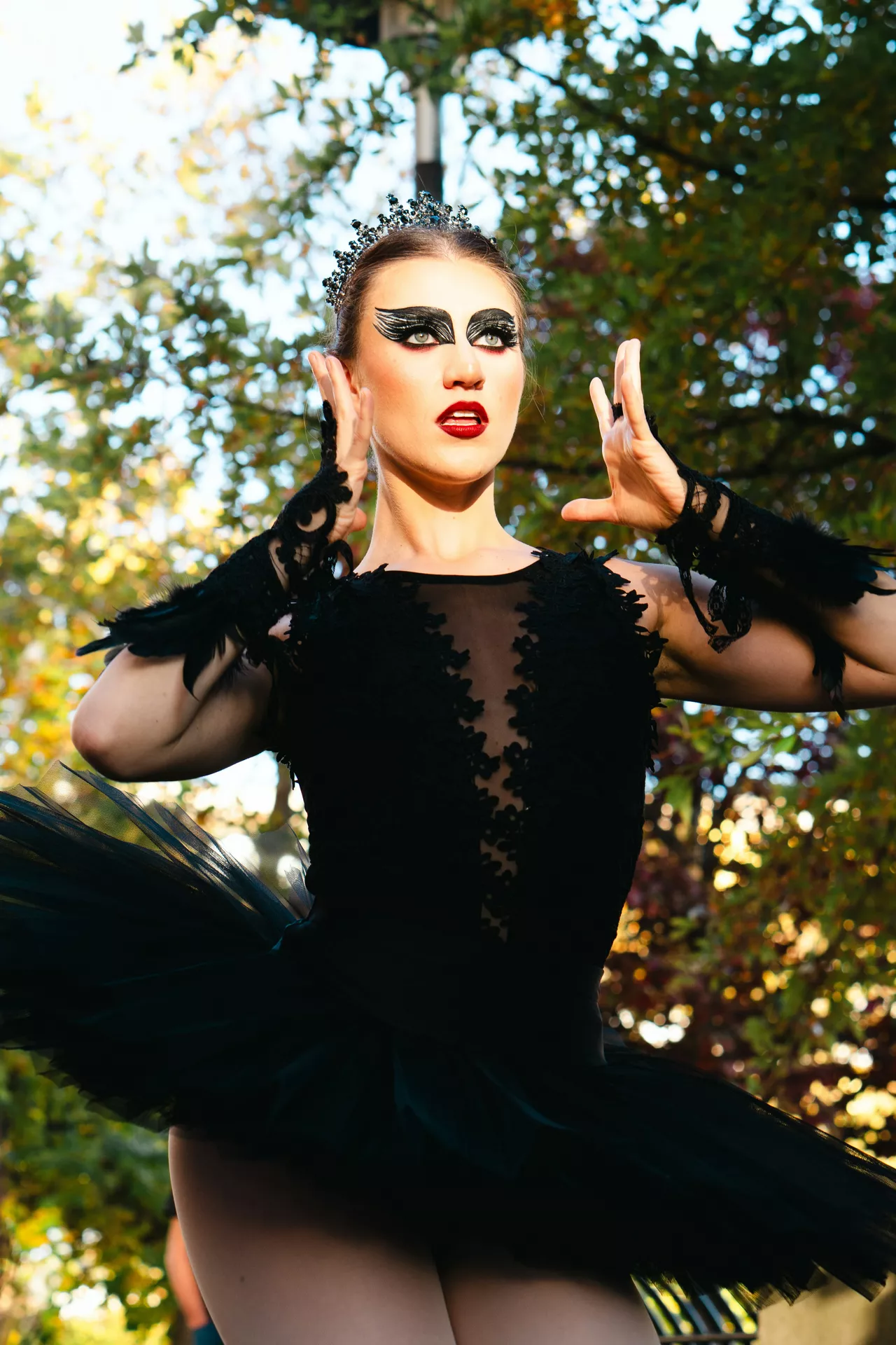 Halloween costume ideas featuring a dramatic black swan look with a black feathered tutu dress, bold winged eye makeup, red lipstick, and an elegant dark crown.