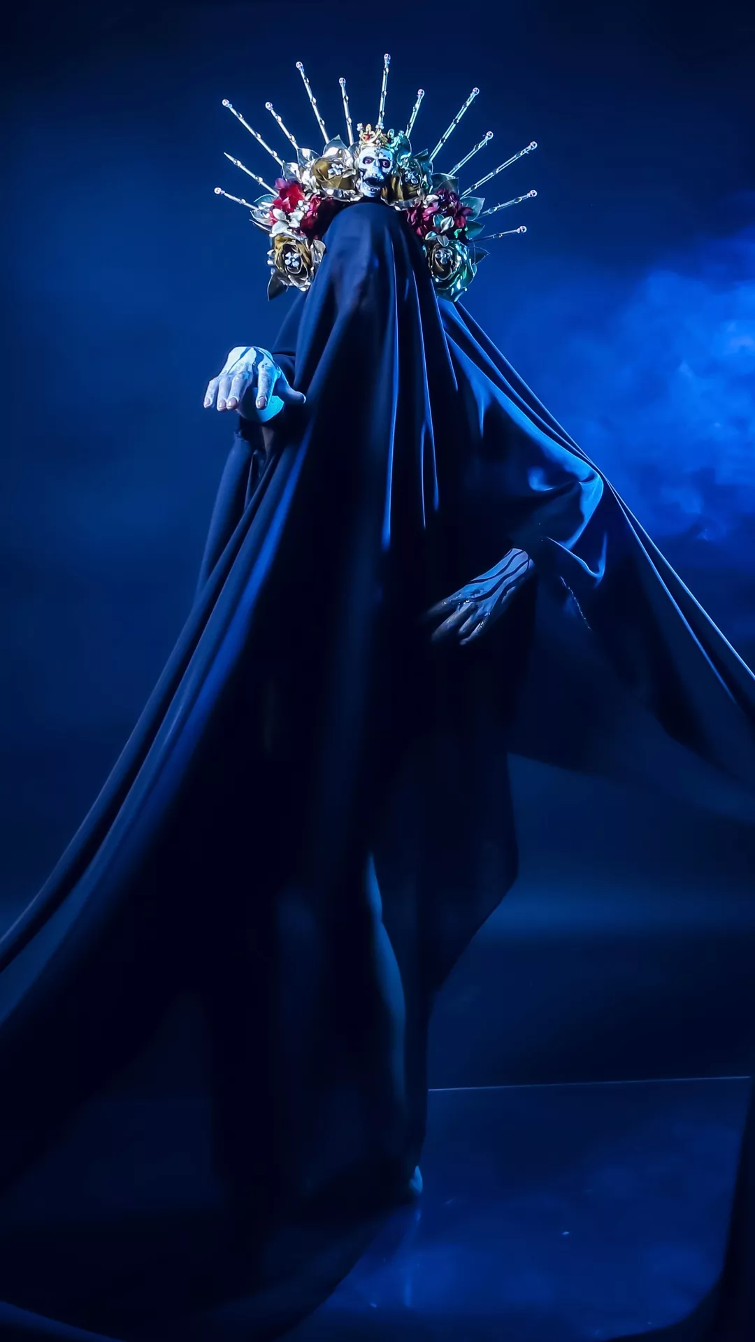 Halloween costume ideas featuring a dark Madonna in a flowing black gown with skeletal makeup, an elaborate floral crown, and a dramatic spiked halo against a smoky blue background.
