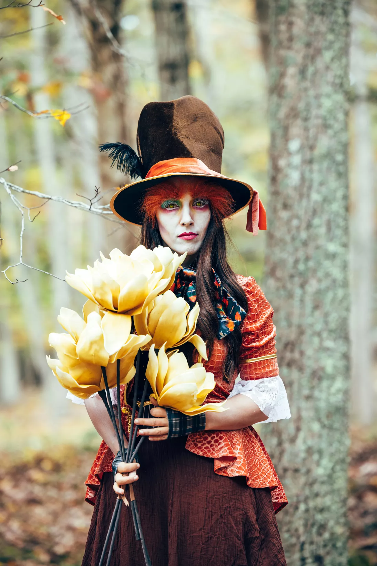 Halloween costume ideas featuring the Mad Hatter from Alice in Wonderland, dressed in a tall brown top hat, orange jacket, colorful makeup, and whimsical accessories while holding oversized yellow flowers in an autumn forest.