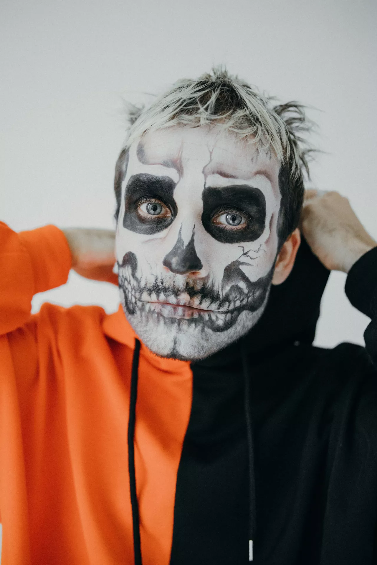 Halloween costume ideas featuring a male skull look with detailed black and white skeleton face paint, dark eye sockets, cracked effects, and a two-tone orange and black hoodie.