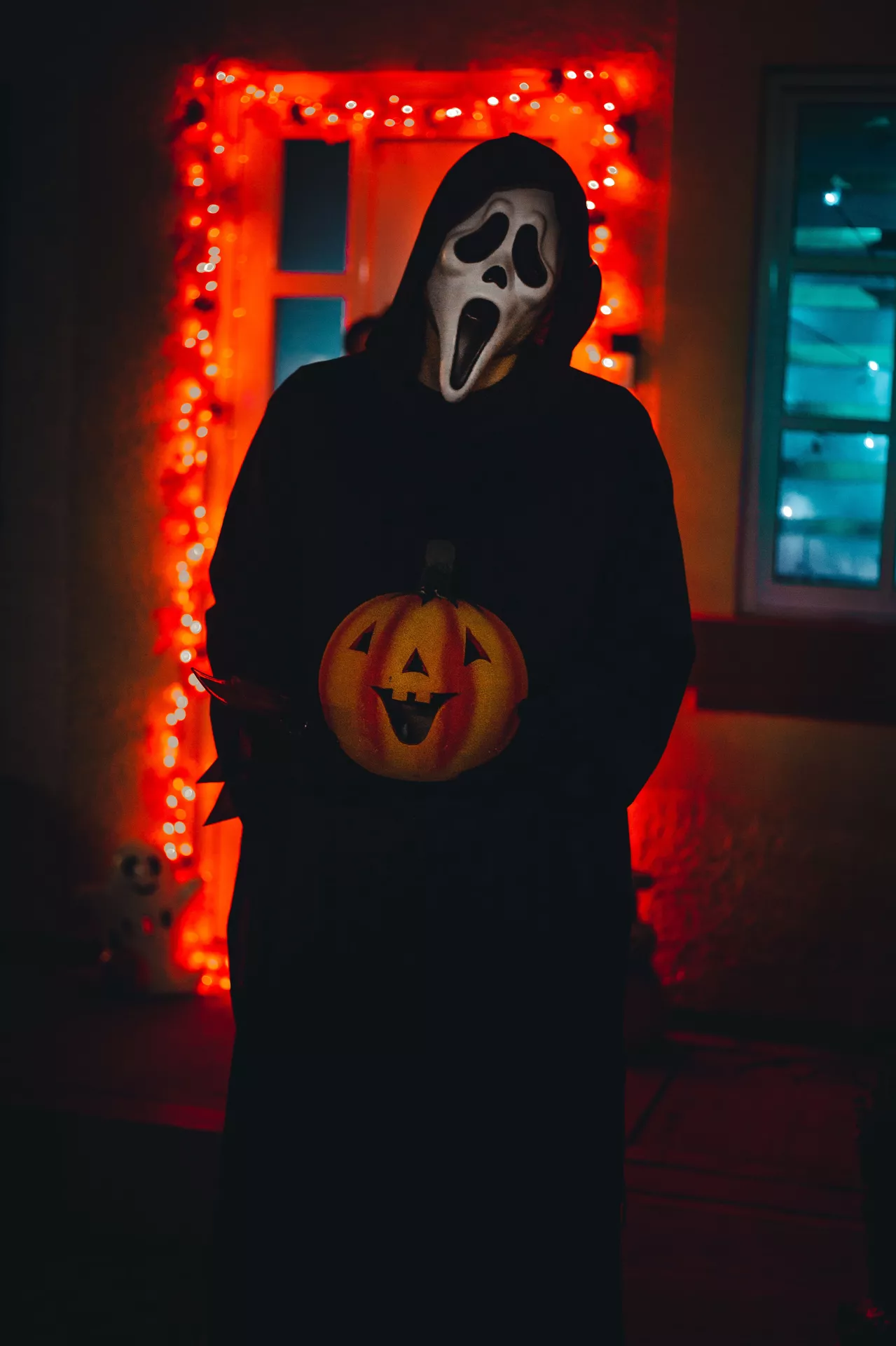 Halloween costume ideas featuring a Scream-inspired character wearing a white ghost mask and long black hooded robe while holding a jack-o-lantern in a dark, orange-lit setting.