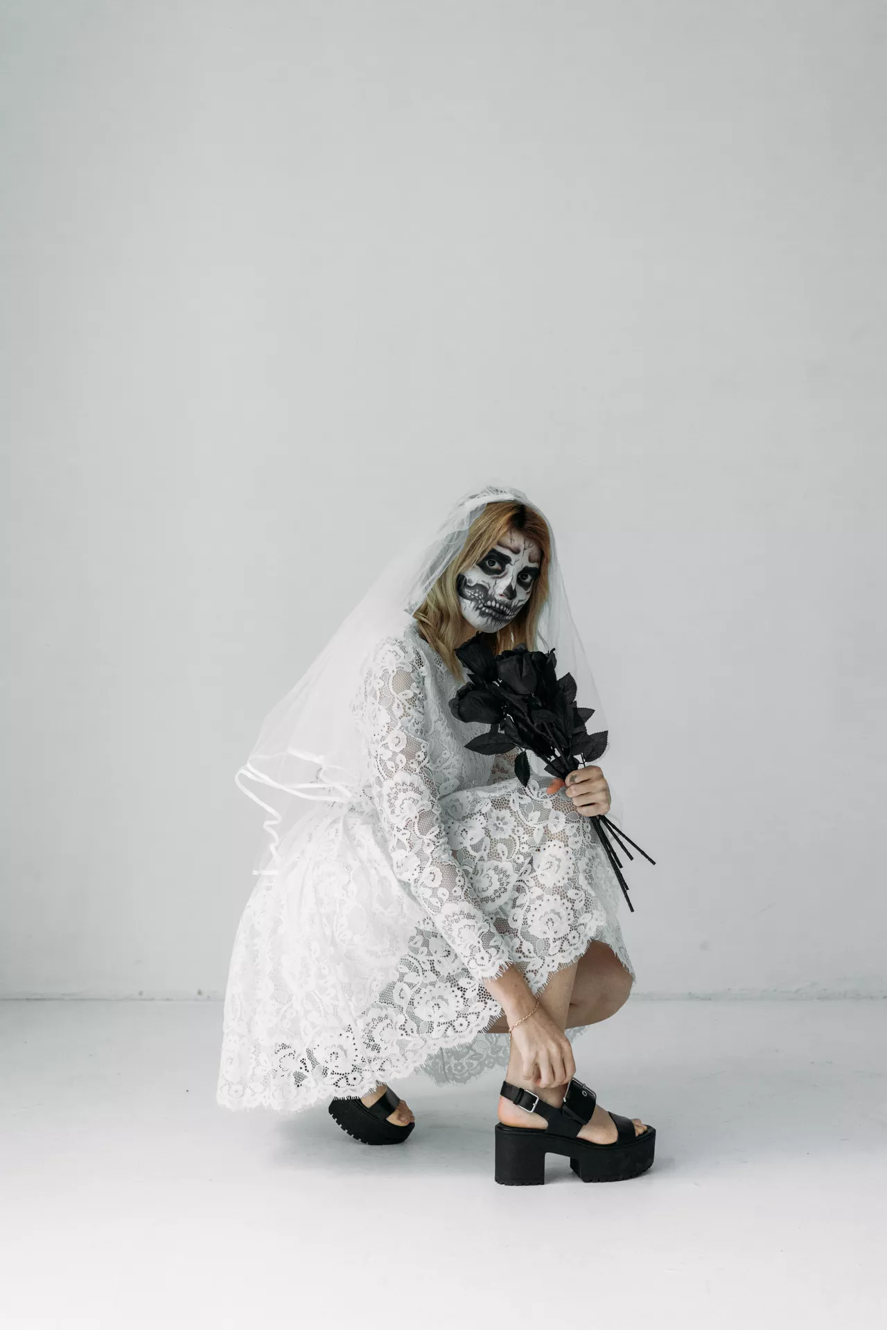Halloween costume ideas featuring a spooky bride in a white lace wedding dress and veil with dramatic skull face paint, black platform shoes, and a bouquet of black roses.