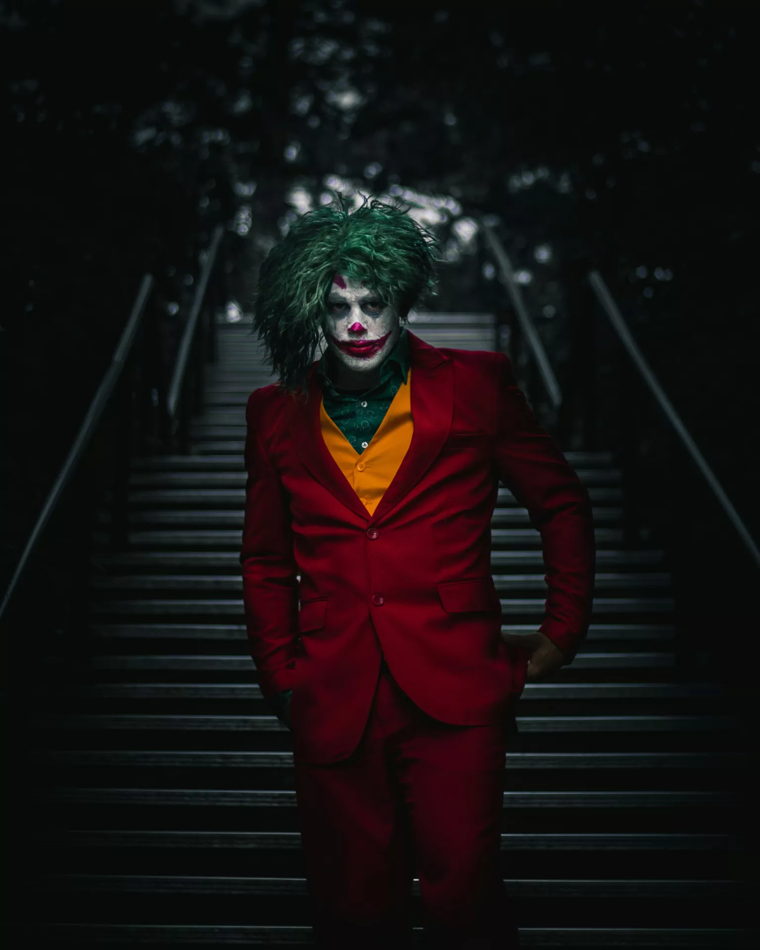 Halloween costume ideas featuring the Joker with green messy hair, white clown makeup, a red suit, yellow vest, and sinister smile posing on a dark staircase.