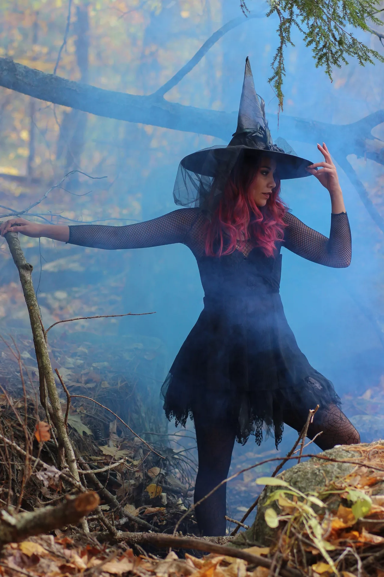 Halloween costume ideas featuring a witch in a black dress, fishnet sleeves, pointed hat, and dark tights posing in a smoky autumn forest.