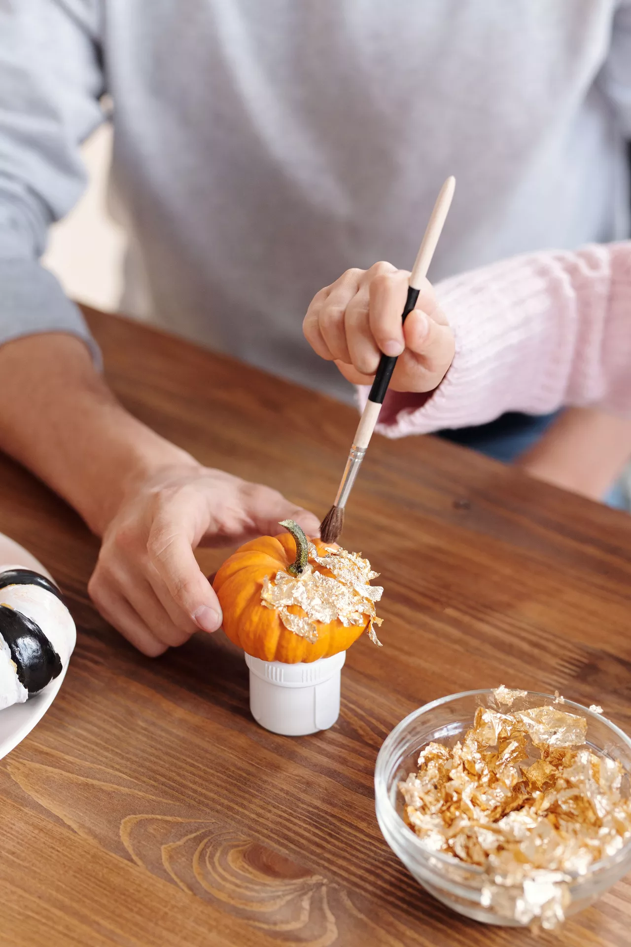 DIY autumn craft ideas featuring Decorating A Pumpkin With Gold Leaf, with a small orange pumpkin being carefully covered in shimmering gold leaf using a paintbrush on a wooden table.