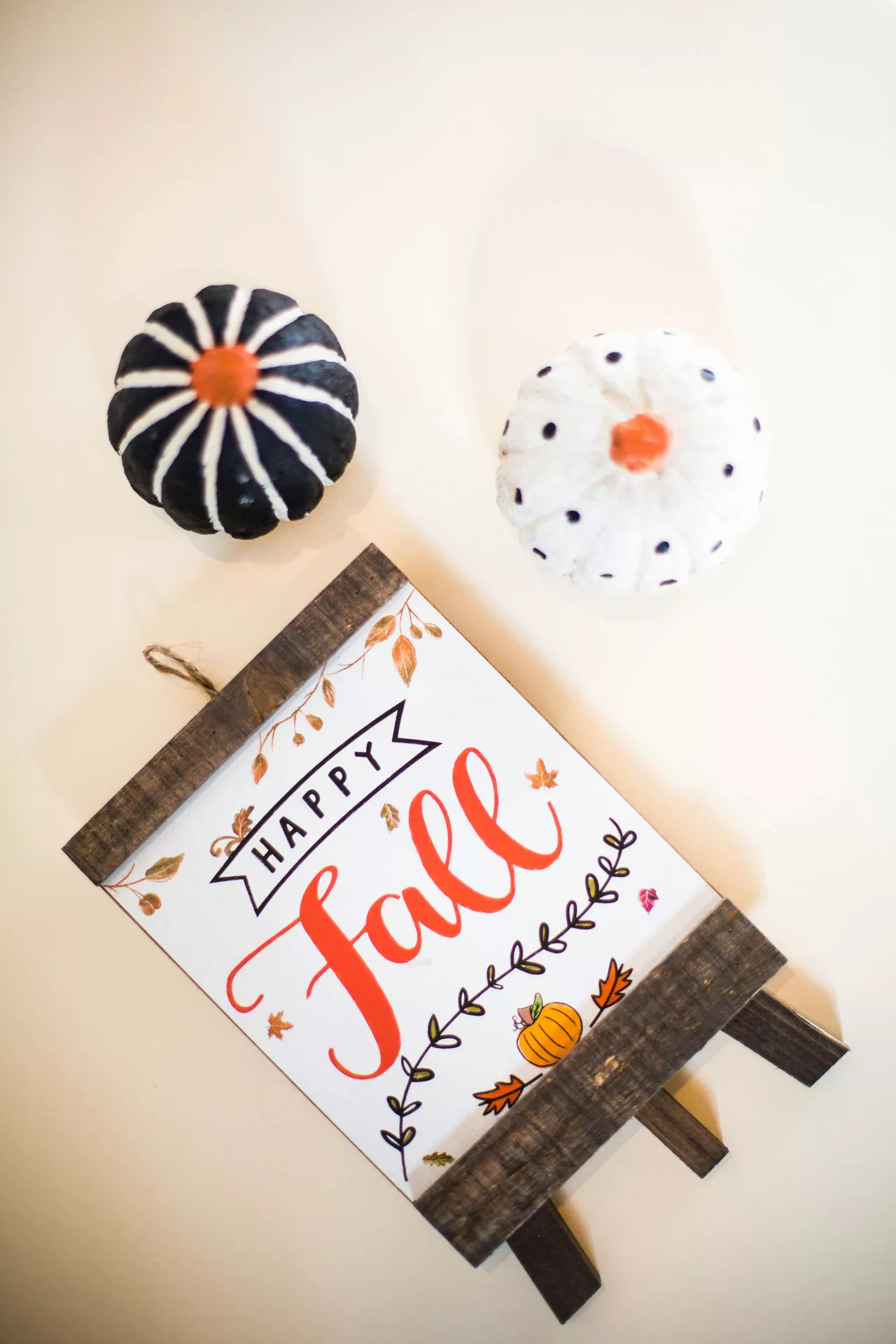 DIY autumn craft ideas featuring fall lettering on a handmade Happy Fall sign decorated with pumpkins, leaves, and botanical details, styled alongside two painted mini pumpkins.
