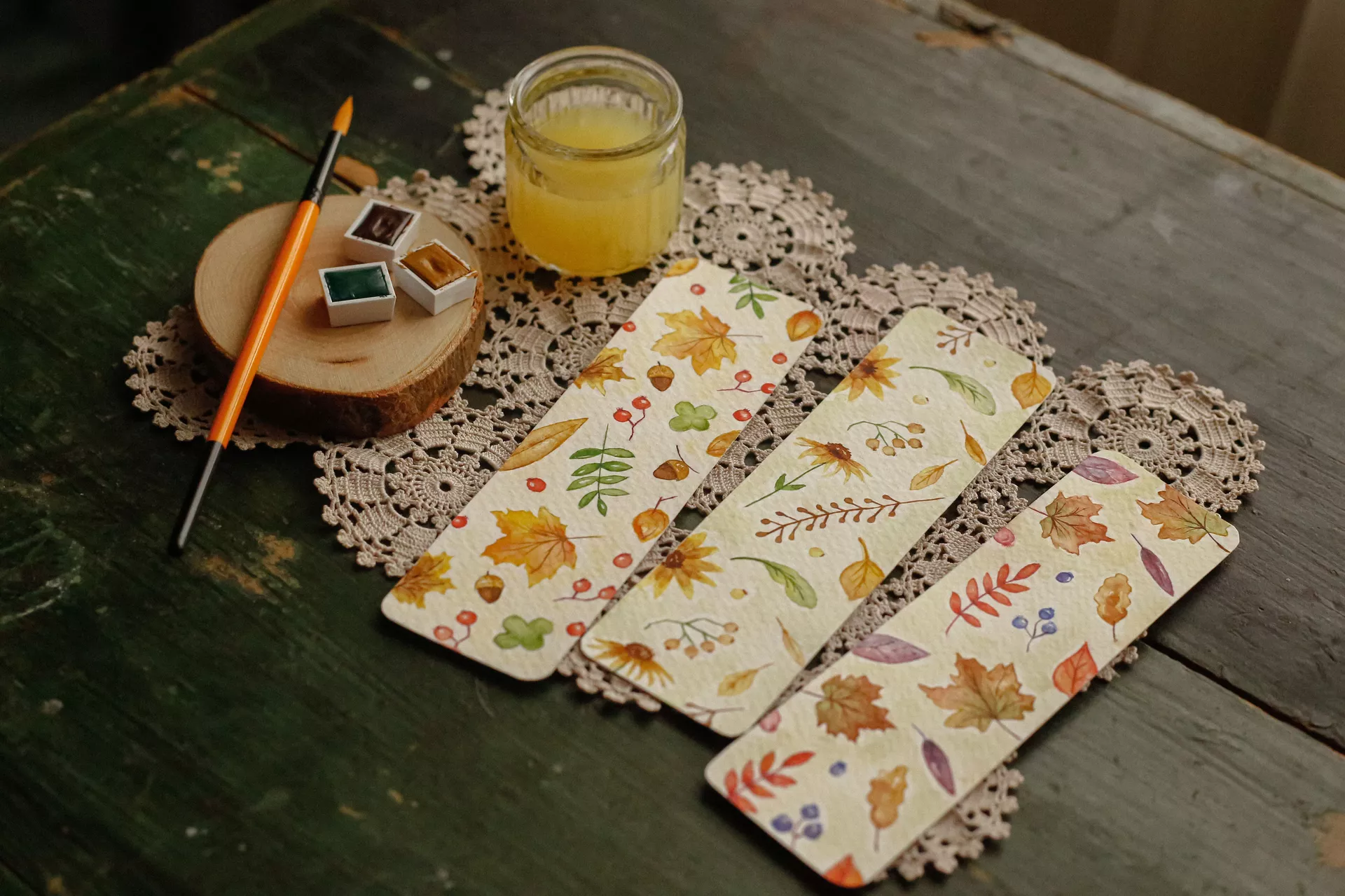 DIY autumn craft ideas featuring painting bookmarks with delicate watercolor leaves, berries, acorns, and botanical motifs in warm fall colors, displayed alongside a paintbrush and watercolor paints.