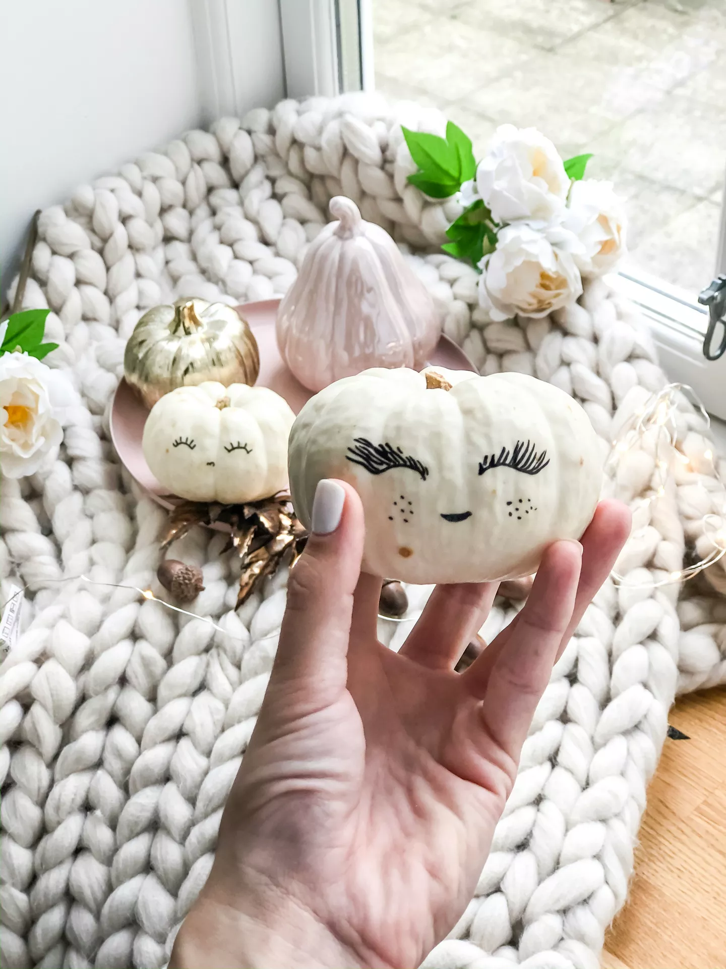 DIY autumn craft ideas featuring pumpkin painting, with a small white pumpkin decorated with a cute sleepy face, rosy cheeks, freckles, and long eyelashes in a cozy fall setting.