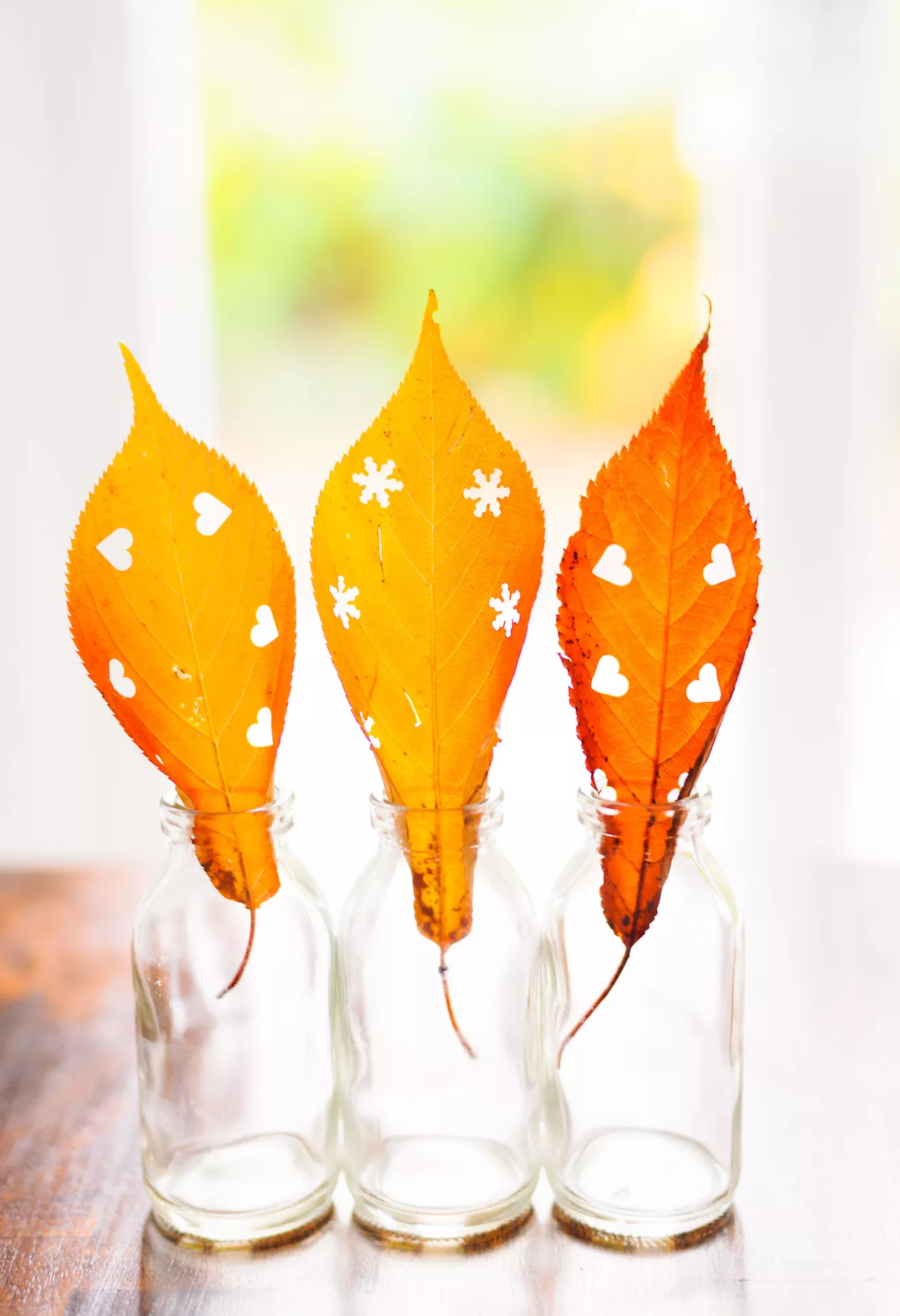 DIY autumn craft ideas featuring punching shapes out of leaves, with three vibrant yellow and orange autumn leaves decorated with heart and snowflake cutouts and displayed in small glass bottles.