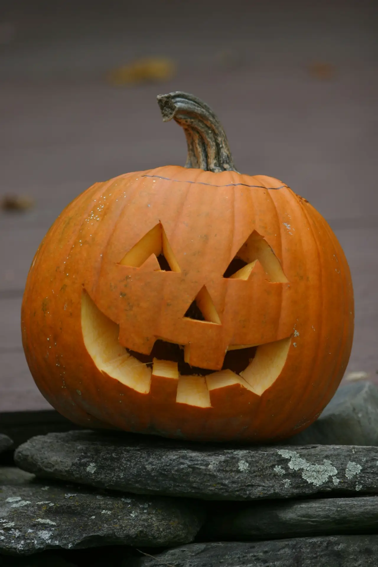Pumpkin carving ideas featuring a classic friendly face with triangular eyes, a small nose, and a wide toothy smile carved into an orange pumpkin displayed outdoors on a stone wall.