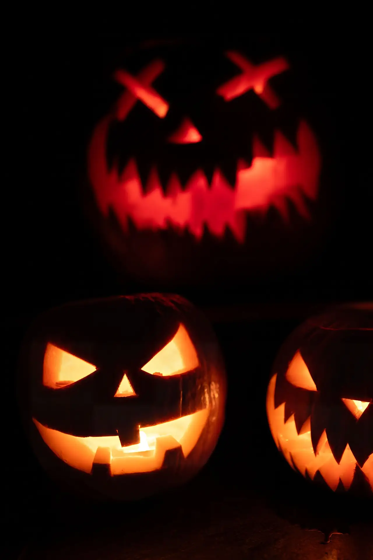 Pumpkin carving ideas featuring three glowing jack-o-lanterns with evil smiling faces, sharp jagged teeth, triangular eyes, and spooky expressions illuminated in the dark.