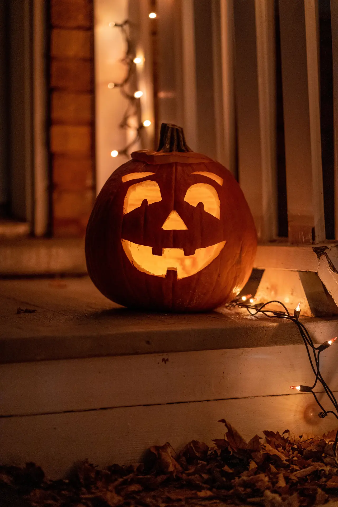 Pumpkin carving ideas featuring a glowing jack-o-lantern with a friendly face with eyebrows, rounded eyes, a triangle nose, and a cheerful toothy smile surrounded by warm string lights.