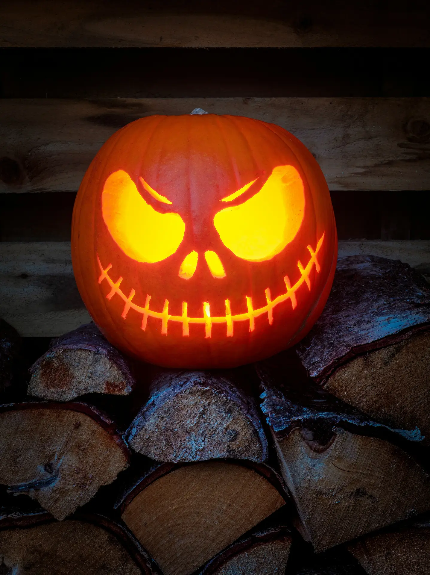 Pumpkin carving ideas featuring a glowing Jack Skellington pumpkin with large menacing eyes, a small skull-like nose, and a wide stitched grin, displayed on stacked firewood for a spooky Halloween look.