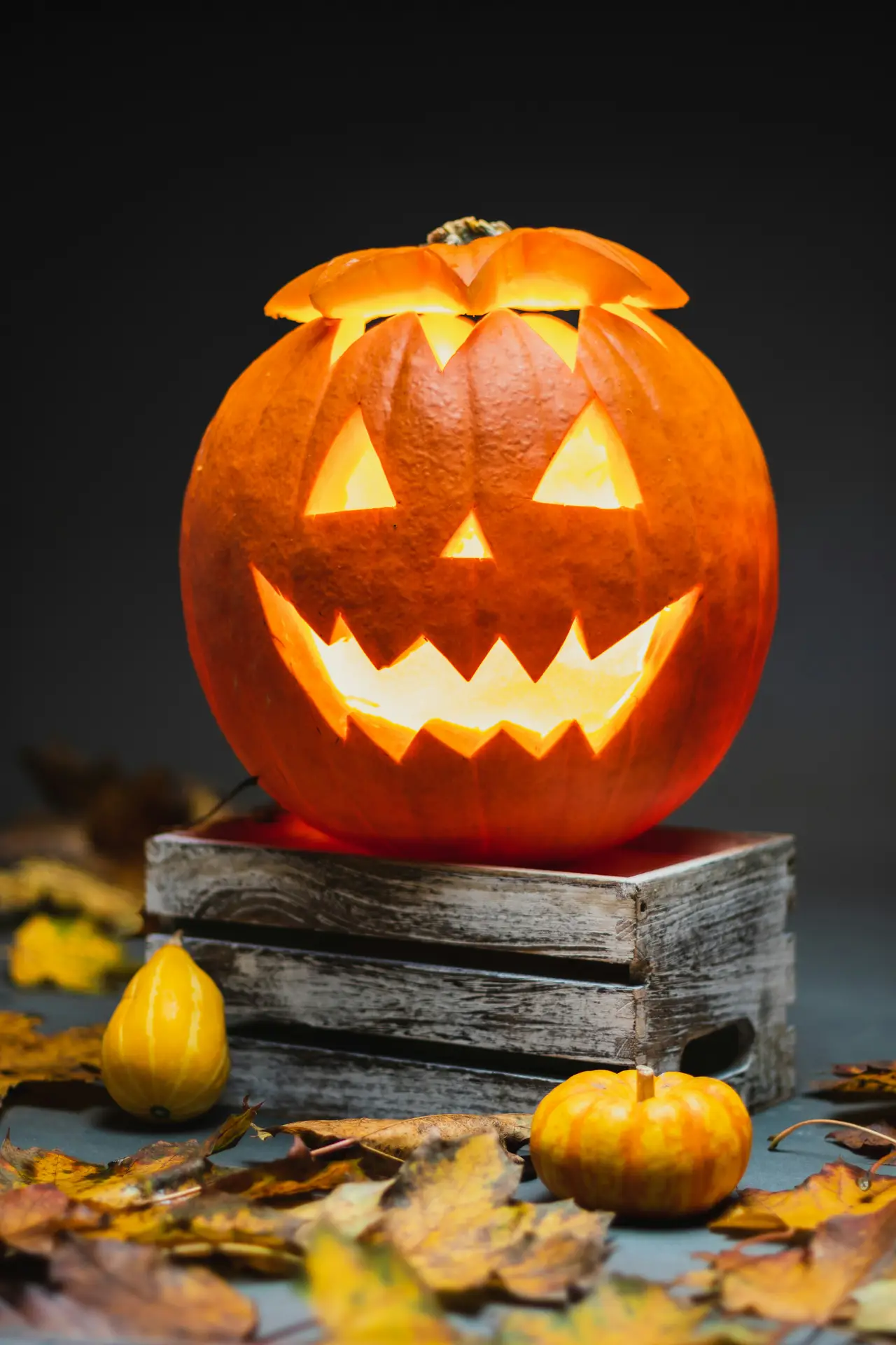 Pumpkin carving ideas featuring a glowing jack-o-lantern with a jagged cheerful face, triangular eyes, a small nose, and a wide zigzag smile, displayed on a rustic wooden crate surrounded by small pumpkins and fall leaves.