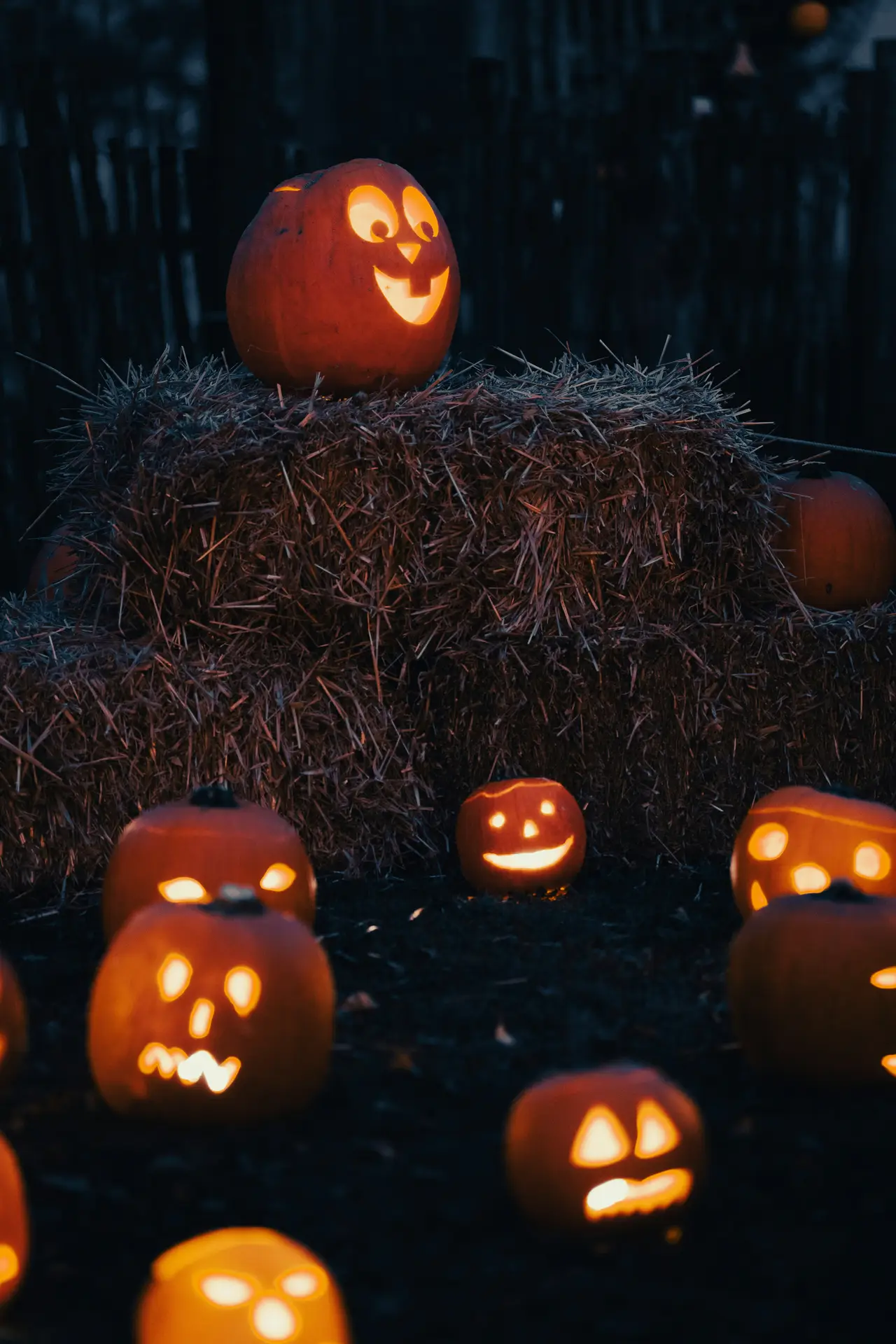 Pumpkin carving ideas featuring a mix of different faces glowing in the dark, with happy, silly, surprised, and spooky jack-o-lanterns arranged around rustic hay bales.